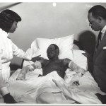 A man in a suit and a woman in a nurse's uniform stand over a hospital bed with a patient at Winslow Hospital.