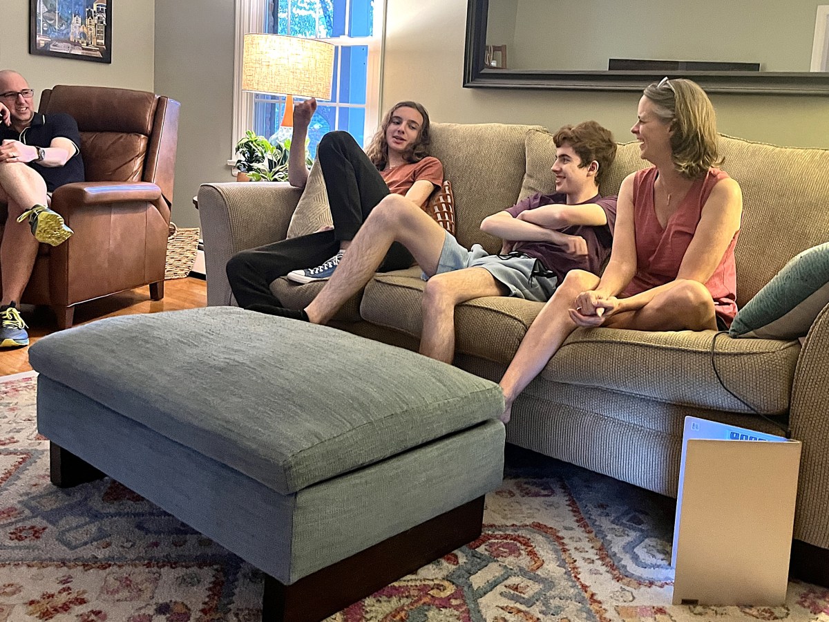 Two teen boys sit on a large couch next to their mother. They speak with their father who sits in a leather chair to the side.