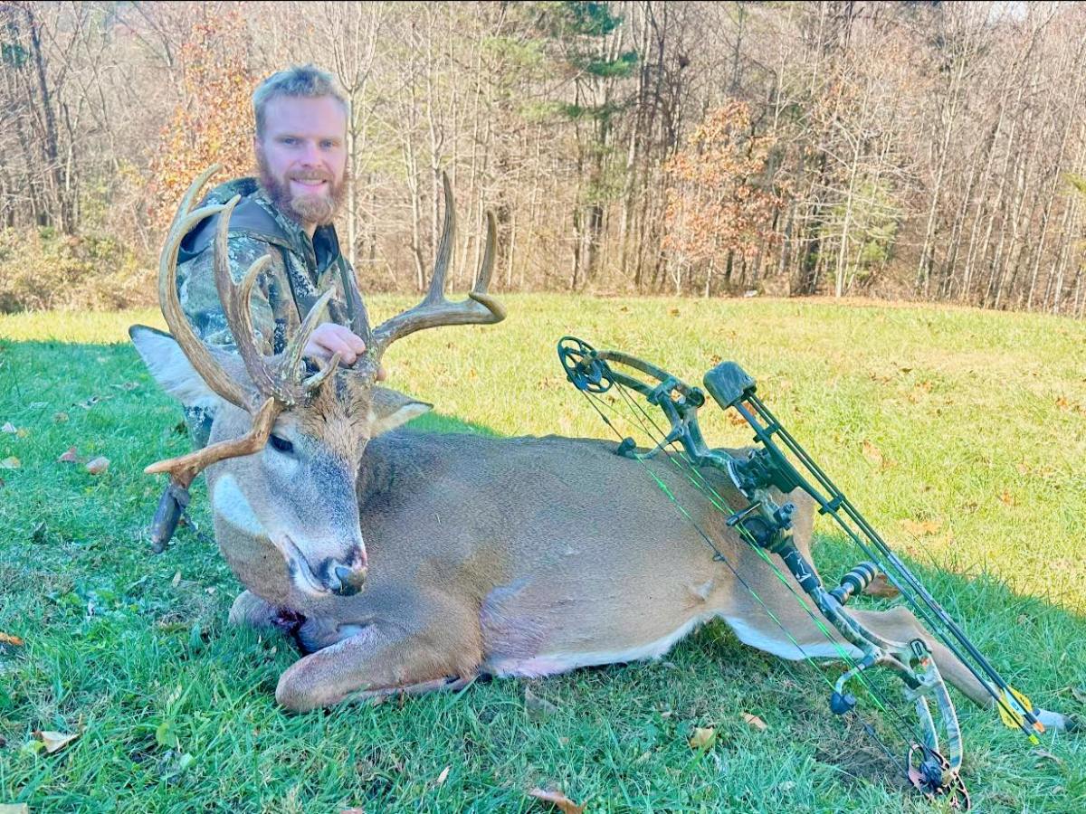 A man in hunting clothes kneels down next to a dead buck in a field.