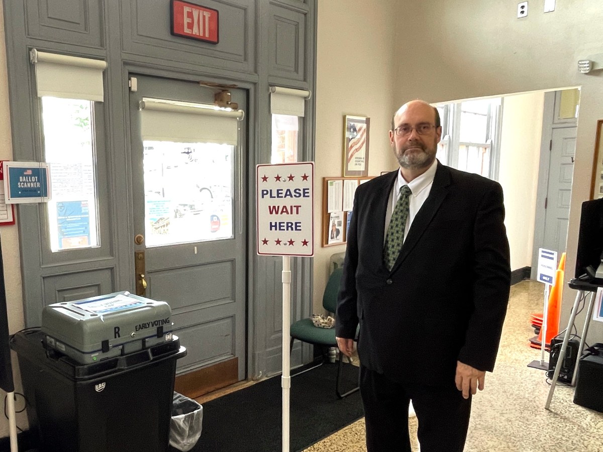 A man in a suit stands by a ballot scanner machine and a sign that reads "Please wait here."